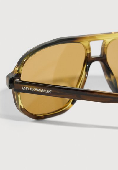Brūnganu toņu Emporio Armani saulesbrilles ar caurspīdīgi brūniem rāmiem, tuvplāns uz kreisās kājiņas, kur redzams logo baltā krāsā.