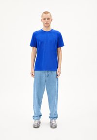 Blaues T-Shirt mit kurzen Ärmeln, kombiniert mit lockeren hellblauen Jeans; Model trägt graue Sneakers; einfarbiger Hintergrund.