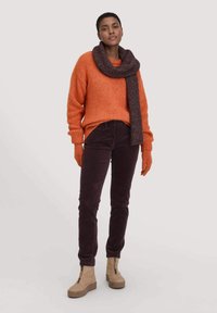 Orange Strickpullover, braune Cordhosen, beigefarbene Plateau-Stiefel und ein dunkler Schal. Die Accessoires umfassen passende orangefarbene Handschuhe.