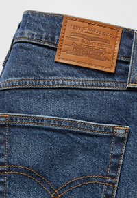 Jeans de mezclilla azul con un parche trasero de cuero marrón que presenta texto en relieve y un gráfico. La costura en hilo de color amarillo dorado añade detalles.