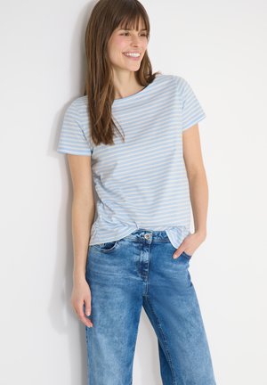 Jonge vrouw die lacht, gekleed in een lichtblauw-wit gestreept shirt met korte mouwen en een blauwe jeans, staand tegen een witte muur.