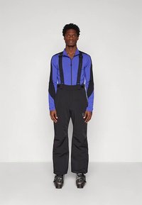 Vyras, vilkintis juodus slidinėjimo kelnes su petnešomis, mėlyną ir juodą užtraukiamą viršutinę dalį bei slidinėjimo batus, stovi prieš paprastą baltą foną.