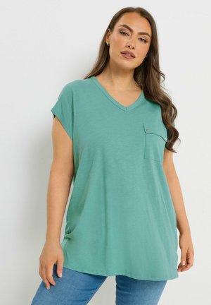 Frau trägt ein locker sitzendes, türkisfarbenes V-Ausschnitt-T-Shirt mit Knopfverschluss an der Tasche und blaue Jeans, steht vor einem schlichten hellen Hintergrund.