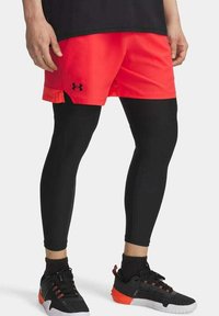 Zwarte compressie leggings gecombineerd met felrode sportshorts. Kenmerken omvatten een logo op de shorts en zwarte sportschoenen met rode accenten.