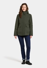 Olivgrön vattentålig jacka med framfickor och huva, i kombination med mörkblå jeans och svarta ankelsboots.