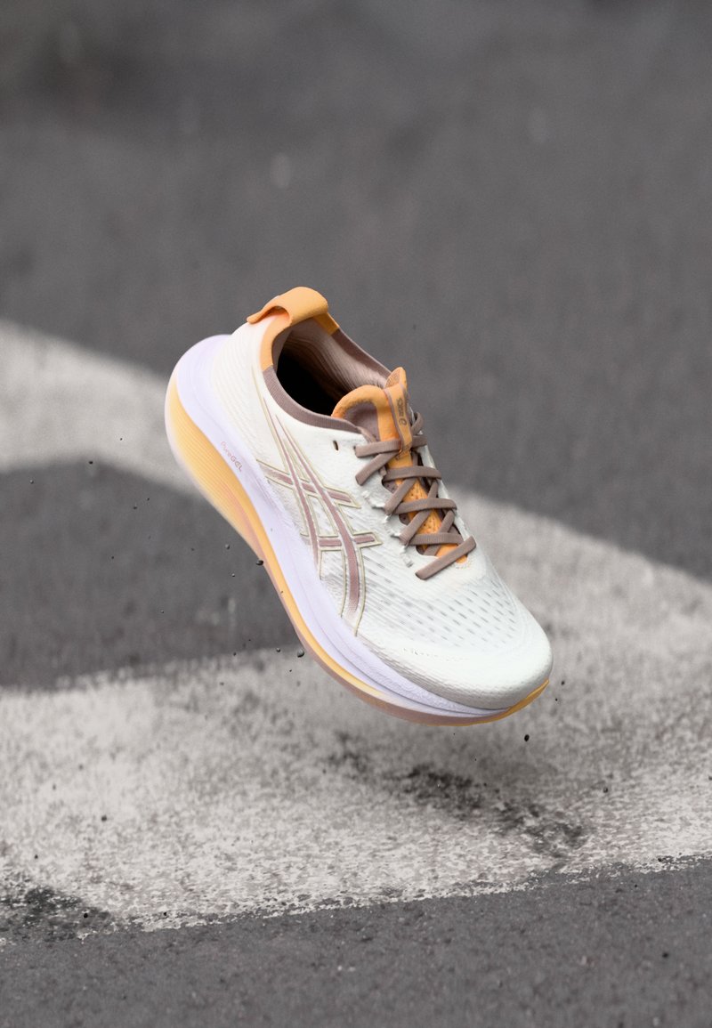 Laufschuh mit einem leichten, strukturierten weißen Obermaterial, orangen Akzenten und einer dicken, gepolsterten Sohle, die ein flexibles Design und eine Schnürung aufweist.