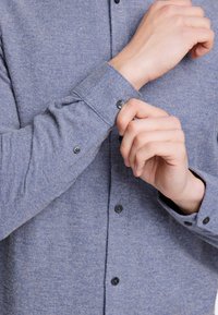 Granatowa koszula z długim rękawem o teksturowanym materiale, z czarnymi guzikami i jednym podwiniętym mankietem dla regulacji.