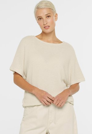 Young woman with short blonde hair wearing a loose, textured short-sleeve beige sweater and light-colored pants, hands clasped.