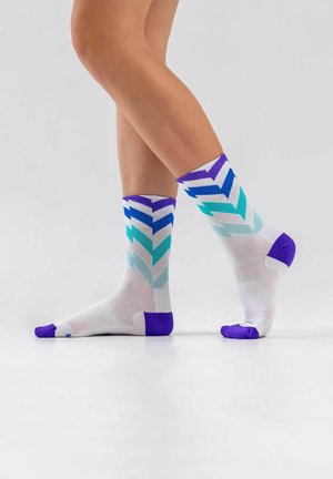 Beine mit weißen knielangen Socken mit lila Zehen und Fersen, die blaue und türkisfarbene Chevron-Muster aufweisen, stehend auf einer weißen Oberfläche.
