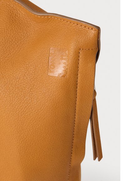 Gros plan d'un coin de sac en cuir beige avec des coutures visibles et un texte en relief sur la surface.