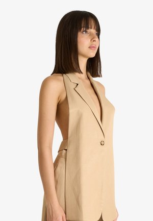 Giovane donna con capelli scuri e lisci indossa un gilet beige senza maniche con scollo a V profondo e un solo bottone, guardando leggermente verso l’alto e a destra.