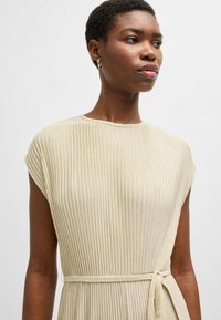 Cream-colored, pleated dress with cap sleeves and a tie belt at the waist; smooth texture and subtle sheen. Close-up view of neckline.