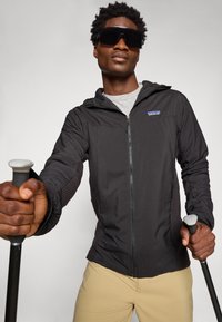 Mees, kes kannab musta lukuga jope ja päikeseprille, hoides suusakeppe, poseerib ühevärvilise heleda tausta ees.