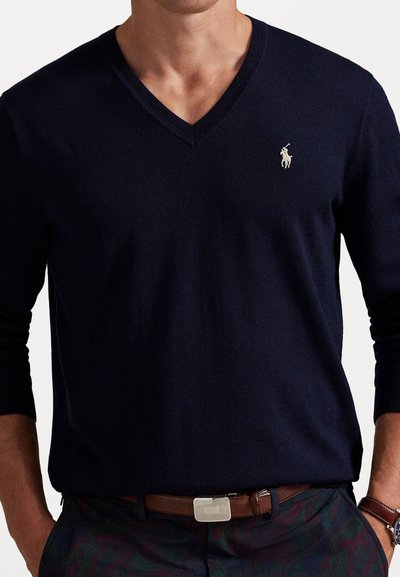 Bărbat îmbrăcat cu un pulover bleu închis cu decolteu în V, având un mic logo brodat cu jucător de polo pe piept, centură maro și pantaloni cu un model paisley.