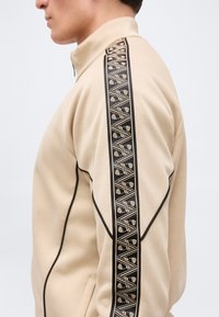 Beige Trainingsjacke mit schwarzem, geometrisch gemustertem Streifen entlang des Ärmels und schwarzer Paspelierung an den Nähten.