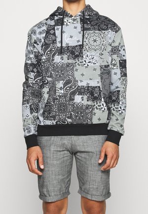 Man draagt een grijze en zwarte patchwork hoodie met paisley patroon, zwarte boorden en trekkoorden, gecombineerd met grijze opgerolde shorts.