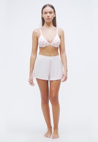 Λευκό σουτιέν με κόκκινα μοτίβα καρδιάς, σε συνδυασμό με ανοιχτό ροζ σορτς. Το ύφασμα είναι απαλό και η εφαρμογή άνετη. Το μοντέλο στέκεται ξυπόλητο.
