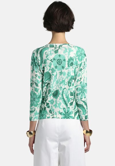 Sieviete ar īsiem tumšiem matiem, kas valkā zaļu ziedu raksta garroku kreklu un baltas bikses, redzama no muguras, ar zelta aproču rokassprādzēm.