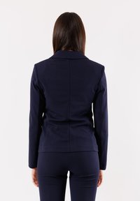 Blazer blu navy, vestibilità sartoriale, spalle strutturate, dettaglio con spacco sul retro, abbinato a pantaloni coordinati in un tessuto liscio. Linee pulite, design minimal.