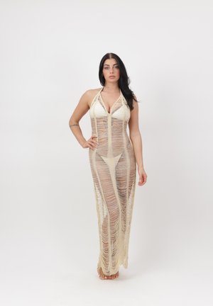 Kvinde med langt mørkt hår iført en beige, revet, ærmeløs maxikjole over en hvid bikini, stående barfodet mod en hvid baggrund.