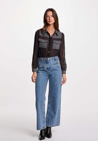 Blusa negra semi-transparente con cuello texturizado y bolsillos, combinada con jeans de pierna ancha de talle alto en azul, llevada con botas negras hasta el tobillo.