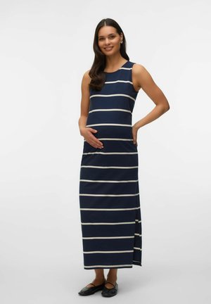 Gravid kvinde med langt mørkt hår iført en ærmeløs maxi-kjole med navyblå og hvide striber og sorte flade sko, smilende, hånd på maven.