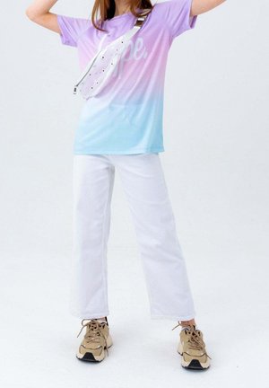 Osoba ubrana w koszulkę z gradientem od purpury do niebieskiego, białe spodnie, beżowe trampki i przezroczystą torbę na ramię na jednolitym tle.