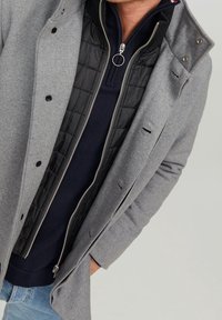 Grå ullkappa med knapplist fram, sidofickor och ett vadderat svart innerfoder med silverdragkedja. Mörkblå pullover under.