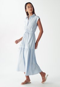 Abito azzurro chiaro con maniche a cappuccio, chiuso da bottoni, con vita elastica, strati arricciati e orlo fluido a lunghezza media. Indossato con sandali piatti.