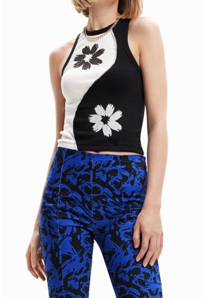 Mujer que lleva una blusa sin mangas negra y blanca con estampados florales contrastantes y pantalones azules con patrón, contra un fondo blanco.