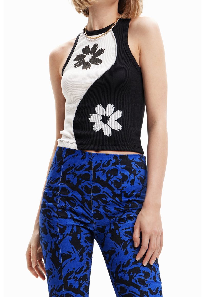 Femme portant un haut sans manches noir et blanc avec des imprimés floraux contrastés et un pantalon bleu à motifs, sur fond blanc.