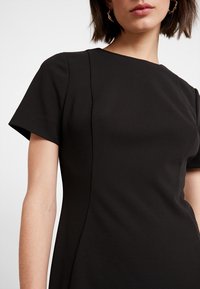 Schwarzes Kurzarmkleid mit lockerer Passform, runder Ausschnitt und glattem, leicht strukturiertem Stoff. Nähte schaffen eine subtile Struktur.