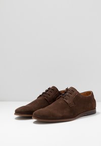 Scarpe eleganti in suede marrone con un design raffinato, punta affusolata e chiusura con lacci. Presentano un interno in pelle liscia e una suola a profilo basso.
