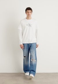 Bijela majica kratkih rukava s sivim logotipom Calvin Klein, nosi se preko bijele košulje dugih rukava. Uparena je s svijetloplavim širokim trapericama s iznošenim efektom.