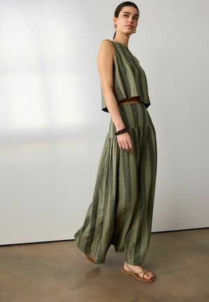 Vrouw loopt binnenshuis en draagt een mouwloze top met groene en donkergroene verticale strepen en een broek met wijde pijpen, gecombineerd met sandalen en armbanden.