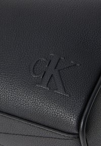 Črna usnjena torba z teksturirano površino, ki ima v dogodku embosiran "ck" logotip. Oblika je pravokotna z čistim šivom in minimalnimi poudarki.