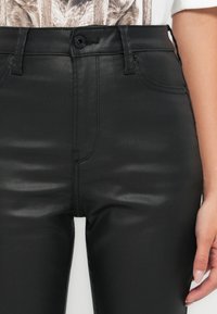 Zwarte neplederen broek met een gladde textuur, hoge taille, voorzakken en een enkele knoopsluiting. Stikstofdetails zichtbaar op de naden.
