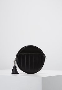 Borsa a tracolla rotonda nera in pelle con cuciture verticali e un tirante a frange sulla cerniera, esposta su un piedistallo bianco.