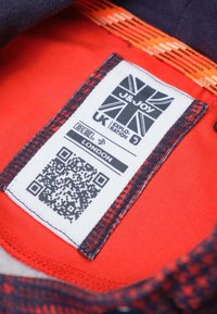 Label van een kledingstuk met een ontwerp van de Britse vlag, een rode stoffen achtergrond, een QR-code en een verstelbare textuurriem in donkerblauw en oranje.