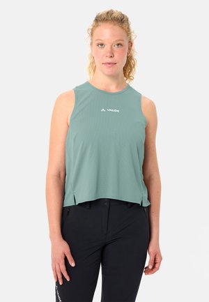 Donna con capelli biondi ricci, che indossa una canottiera teal di Vaude e pantaloni neri, in piedi davanti a uno sfondo chiaro e semplice.