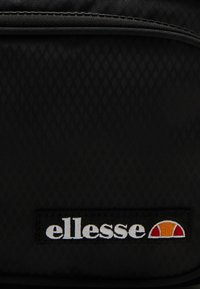 Černý batoh s texturovaným diamantovým vzorem, který je zdobený obdélníkovou výšivkou s názvem značky "ellesse" v bílé, červené a oranžové barvě.