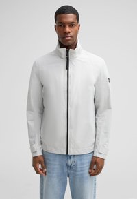 Hellgraue Jacke mit hohem Kragen, schwarzem Reißverschluss und elastischen Bündchen, die eine glatte Stoffstruktur und ein minimalistisches Design aufweist.