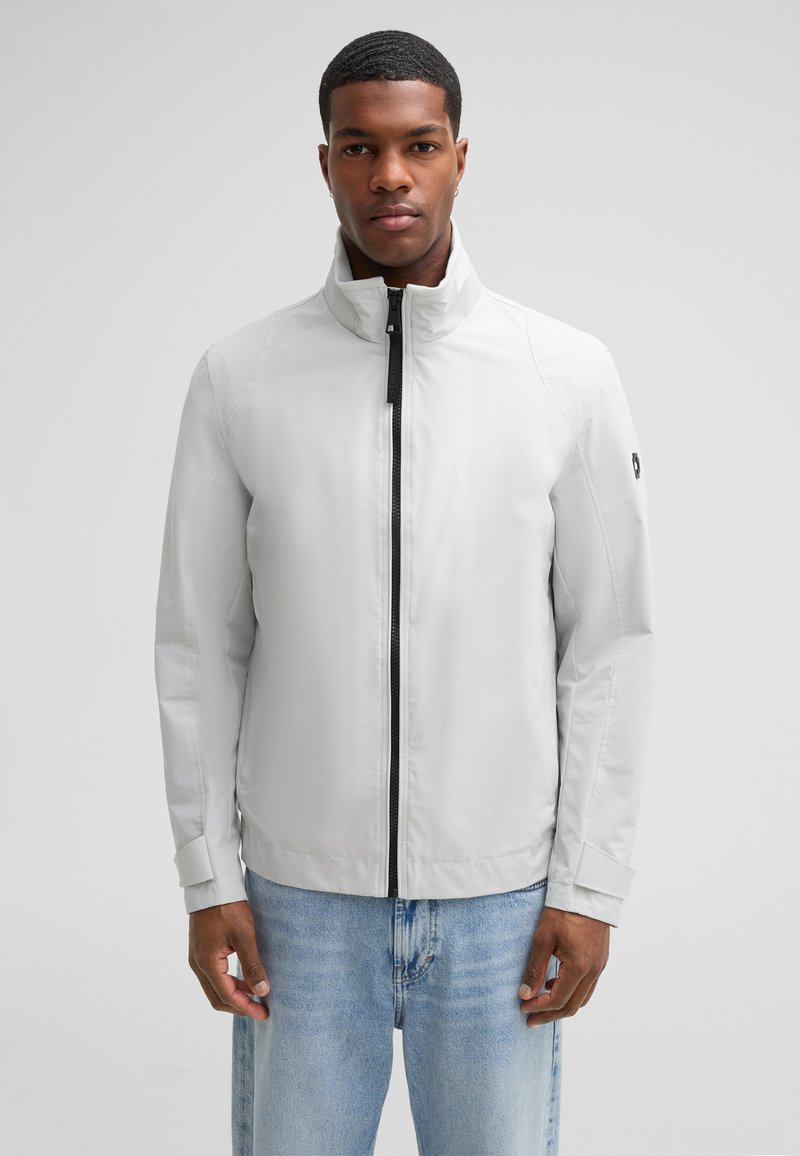 Hellgraue Jacke mit hohem Kragen, schwarzem Reißverschluss und elastischen Bündchen, die eine glatte Stoffstruktur und ein minimalistisches Design aufweist.