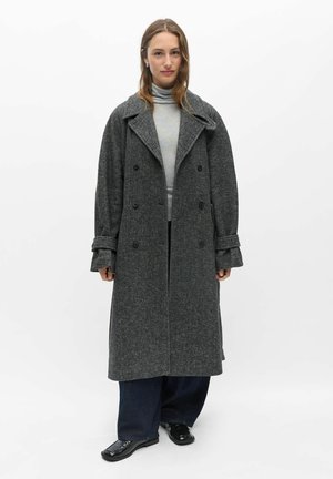 Femme portant un long manteau gris foncé à double boutonnage sur un pull à col roulé gris clair, pantalon large foncé et chaussures noires sur fond blanc.