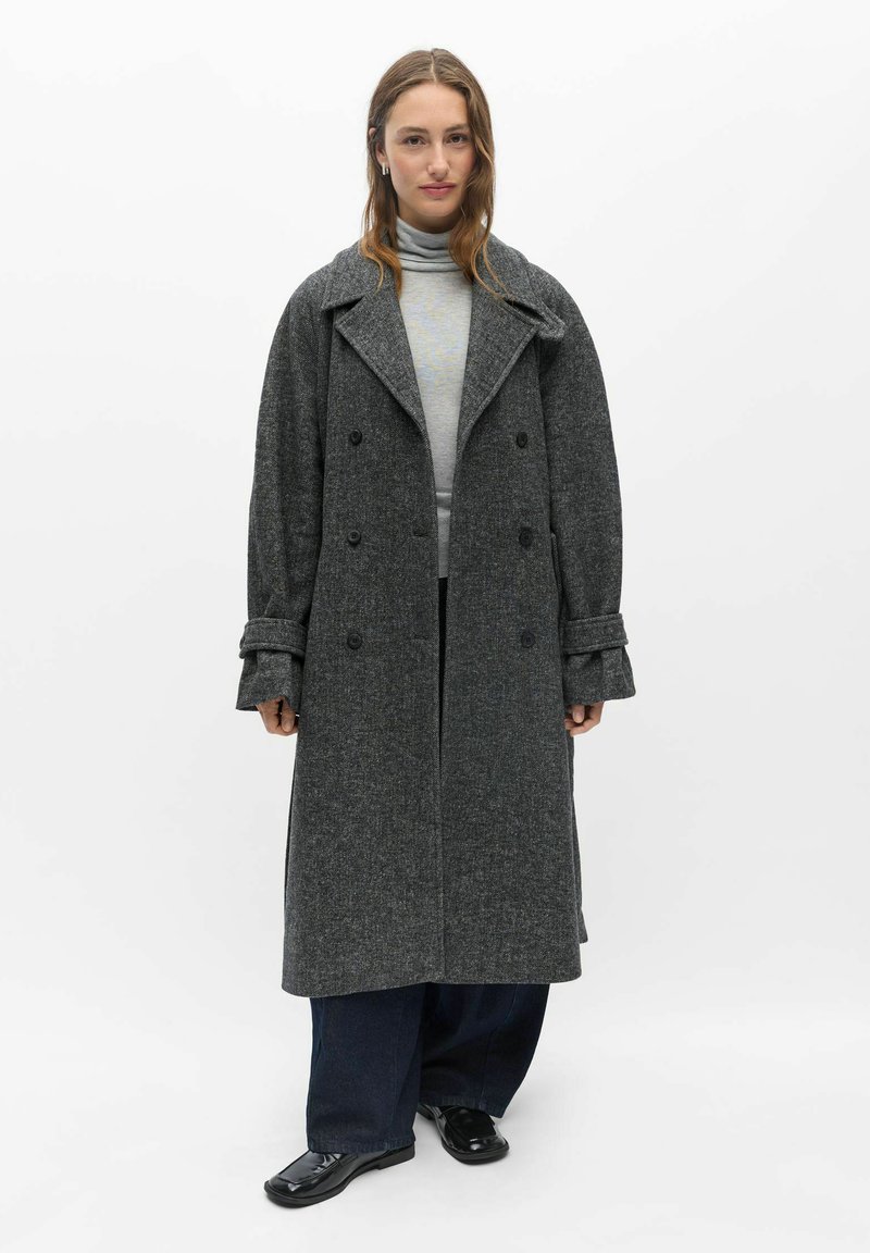 Femme portant un long manteau gris foncé à double boutonnage sur un pull à col roulé gris clair, pantalon large foncé et chaussures noires sur fond blanc.