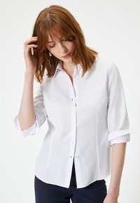 Hvid skjorte med knap, slim fit, lange ærmer op-rullet til albuerne, glat tekstur, subtil glans, buet hem, klassisk krave, fem knapper.