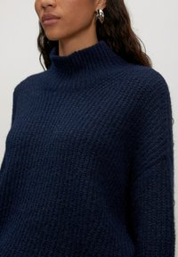 Navy gestrickter Pullover mit hohem, geripptem Kragen und lockerer Passform. Verfügt über ein strukturiertes Muster und ein weiches, wollähnliches Material.