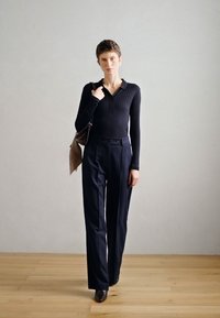 Top de manga longa em malha riscada na cor azul-marinho, combinado com calças largas de risca de giz. O modelo segura uma bolsa de ombro castanha e usa sapatos de salto.