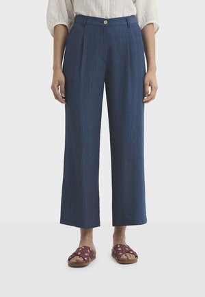 Femme portant un pantalon bleu marine taille haute à jambes larges, une chemise blanche boutonnée rentrée et des sandales rouges à bout ouvert avec des brides à motifs.