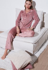 Pinky velur pidžama set s uzorcima mjeseca i zvijezda, koji se sastoji od dugmetne bluze i labavih hlača, uparenih s svijetlim plišastim jastucima.